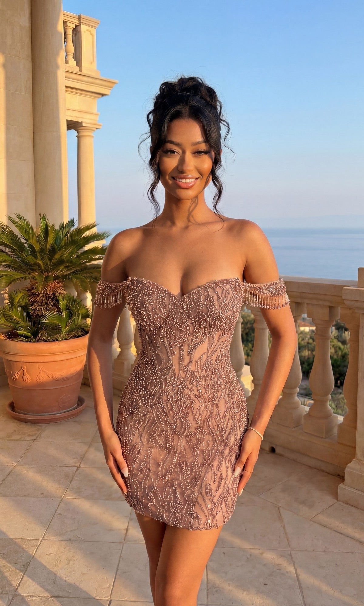 A woman in a rose gold Beaded Glitter Hoco Dress: PromGirl Exclusive 250133 stands on a balcony with a potted palm, columns, and an ocean view in the background.